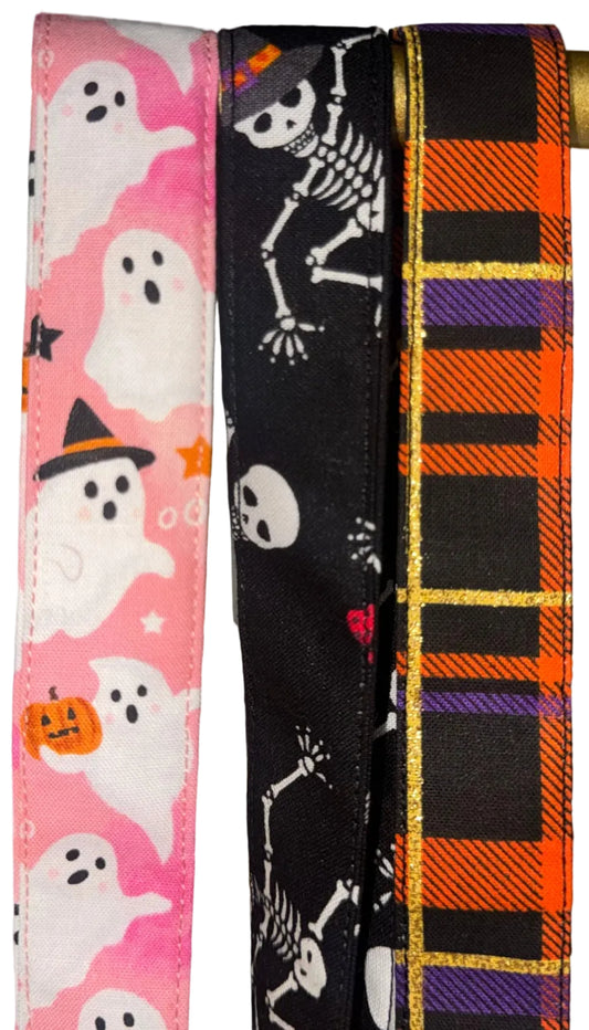 Three Halloween-themed lanyards hanging from a gold rod: a pink ribbon with white ghosts, a black ribbon with white skeleton motifs, and an orange-and-purple plaid ribbon with gold stripes.