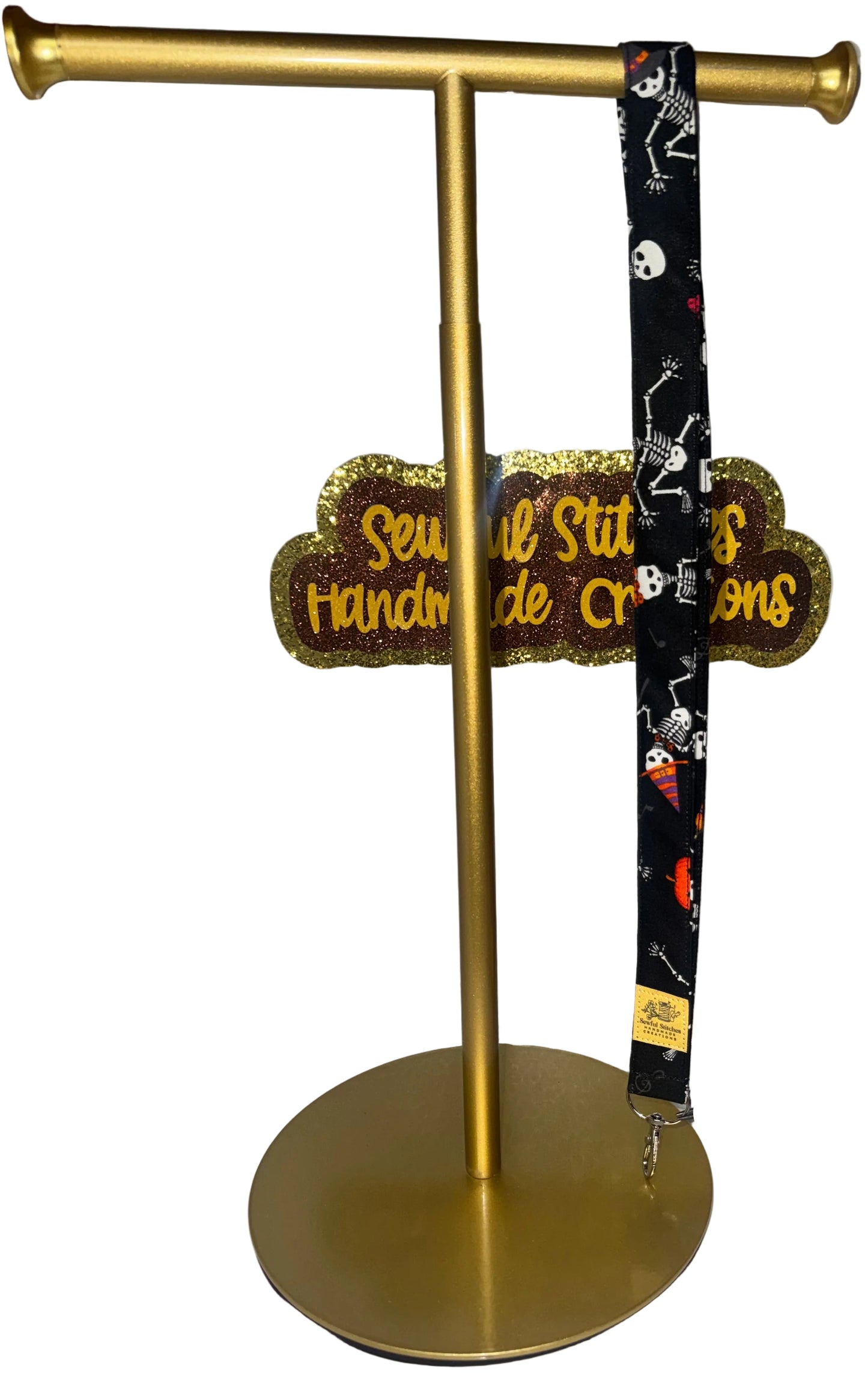 A black Halloween-themed lanyard with a white skeleton print and orange accents is displayed on a gold stand, . A sign in the background reads 'Sewful Stitches Handmade Creations'.