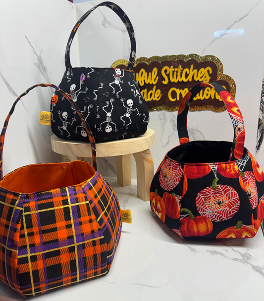 Three handmade pumpkin shaped handbags with Halloween-themed patterns are displayed on a white marble surface. One bag features a black background with white skeleton dancers, another has an orange, purple, and yellow plaid pattern, and the third shows a black background with orange pumpkins and red roses.