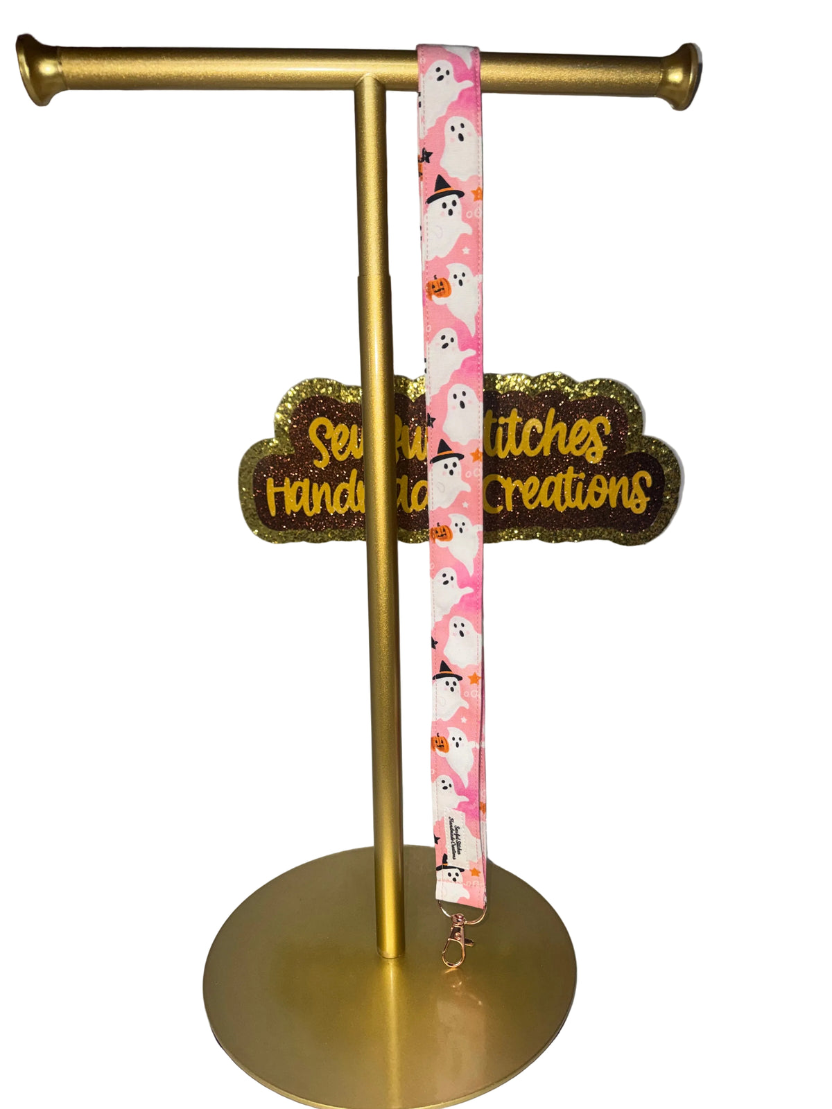 A pink fabric lanyard with a white ghost and black witch hat pattern, hanging from a gold metal display stand.