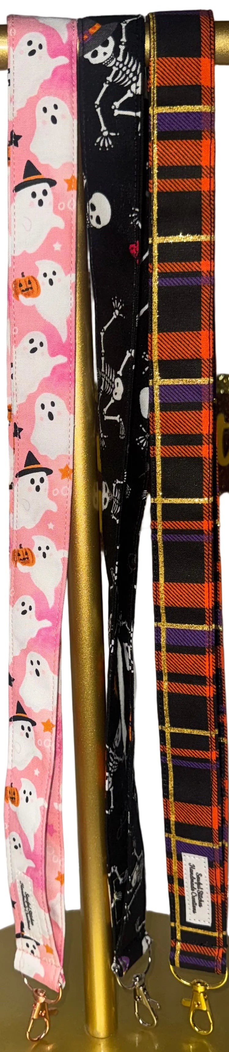 Three lanyards displayed on a gold stand: one pink with white ghosts and orange pumpkin motifs, one black with white skulls and orange accents, and one orange and black plaid.