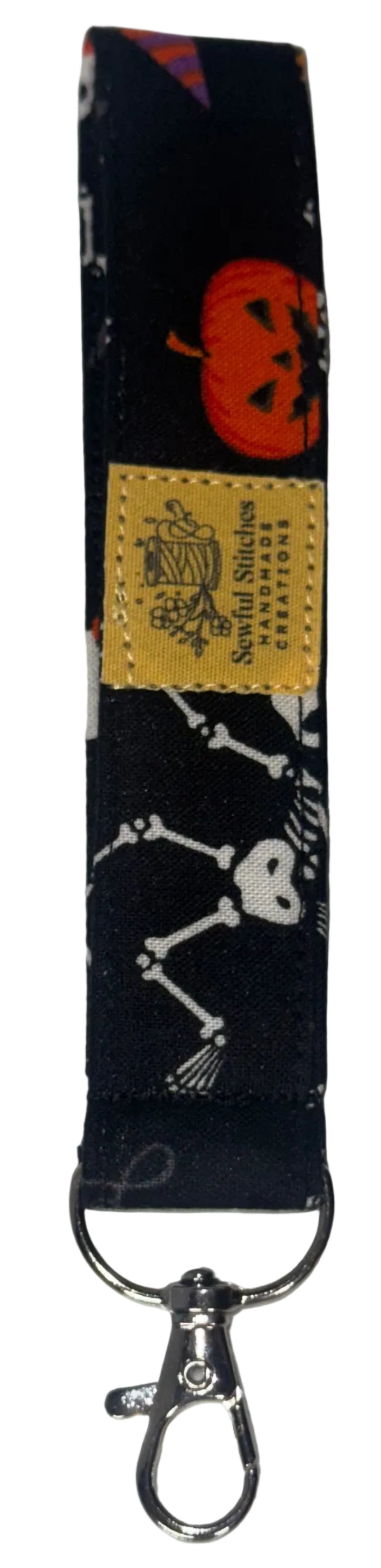 A fabric wristlet keychain strap with a Halloween print, featuring a jack-o'-lantern, skeleton, and broomstick pattern on a dark background, with a yellow brand label and a silver metal clasp, shown on a white surface.