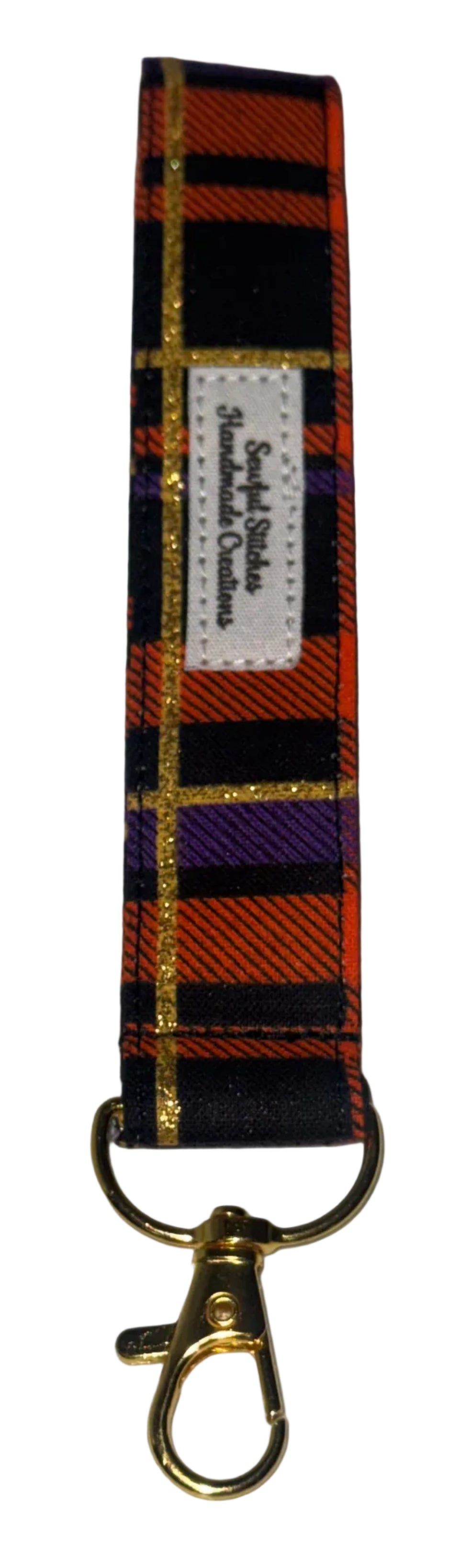 A fabric keychain strap featuring a orange, black, and purple tartan plaid pattern with gold glittery stripes, finished with a white brand label and a gold-tone lobster clasp, displayed on a white  surface.