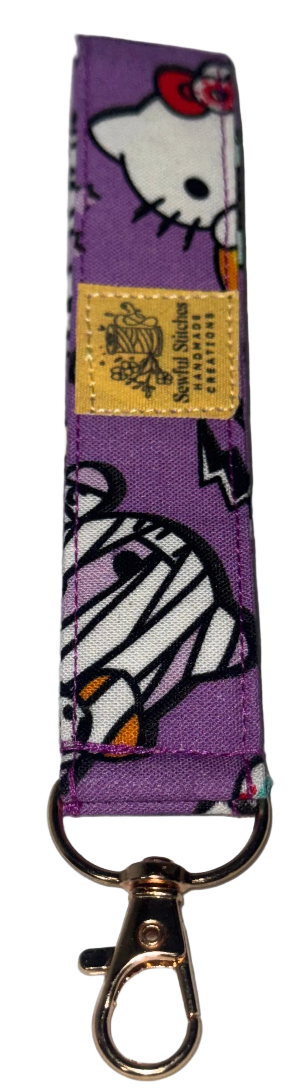 A purple fabric keychain strap featuring mummy print, with a gold-tone metal swivel clasp at the bottom and a small yellow brand label.