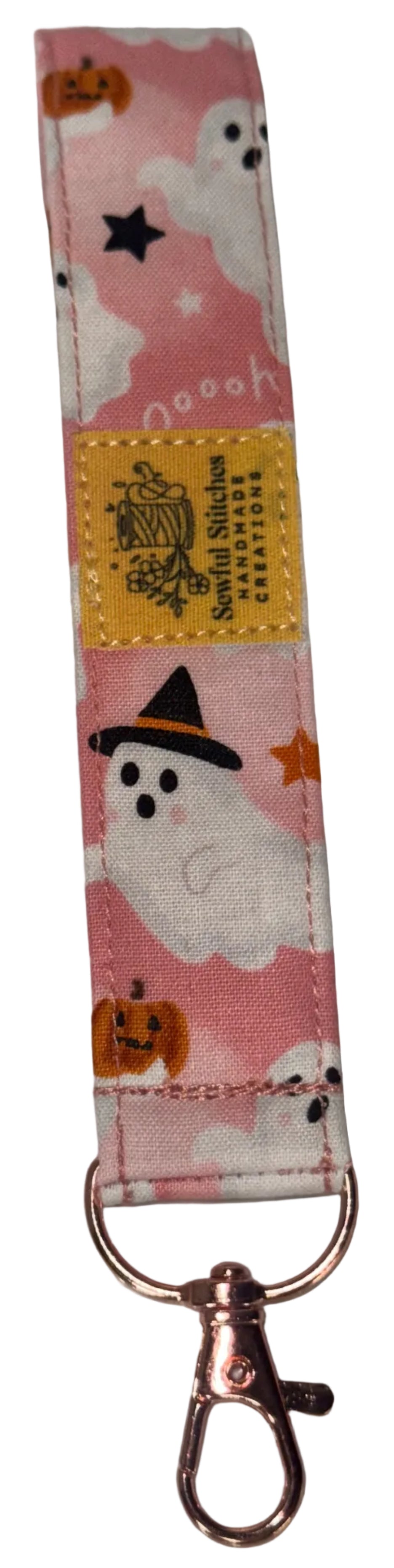 A pink Halloween-themed fabric wrist strap keychain featuring cartoon ghosts, pumpkins, and stars, with a yellow brand label and a rose gold metal clasp, resting on a white surface.