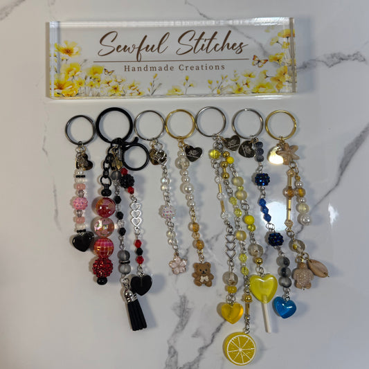Collection of keychains with various charms on a marble surface, branded 'Sewful Stitches'.